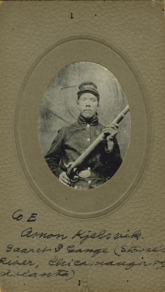 Waist-up oval studio portrait of Anon Kjellsvig [as written on the published Roster of Wisconsin Volunteers, Volume 1; also written as Amon Kjellsvig on the Regimental Descriptive Rolls, Volume 20, and written as Amon Kjelsvik on card backing of portrait], a private in Company E, 15th Wisconsin Infantry, in uniform with a musket across his lap. The following information was obtained from the Regimental and Descriptive Rolls, Volume 20: He resided in York, Wisconsin. On November 29, 1861, he enlisted in Green County, Wisconsin and was mustered into service in Madison, Wisconsin on December 08, 1861, at the age of 22. He was captured and wounded slightly during the battle at Stone River, Tennessee. He was gained from missing in action and found to be a paroled prisoner of war at St. Louis, Missouri. He returned to active duty on June 9, 1863. He was wounded in the ankle during the Battle of Chickamauga, and was sent to a hospital in Nashville, Tennessee.  On August 12, 1864 he was accidentally severely wounded in his hand near Atlanta, Georgia. He mustered out with Company E on December 20, 1864, at Chattanooga, Tennessee.