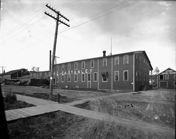 Several buildings in the complex of the F.G. and C.A. Stanley Manufacturing Plant. The largest building is labeled "F.G. and C.A. Stanley." A smaller building to the right is labeled "Storm Sash and Doors." A plank sidewalk and utility pole are in the foreground. Two horses hitched to a wagon stand in the background.