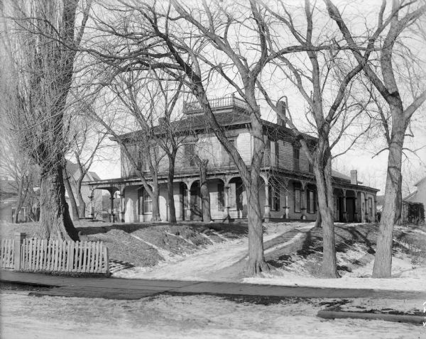 View from road of exterior of house on the H.S. Allen homestead. The house has a widow's walk and a wrap-around porch. There are many large trees on the property and snow is on the ground.