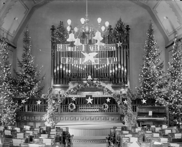 Interior view of the First Presbyterian Church chancel decorated for Christmas. There are several decorated Christmas trees, an arch and crosses covered with evergreen boughs, and paper stars and bells. A large Stere ad Turner pipe organ is on the back wall of the church and bears a large banner reading "GLORY TO GOD IN THE HIGHEST / ON EARTH PEACE AND GOOD WILL." Below and in front of the aisle are an array of music stands. A large chandelier hangs from the ceiling, and an upright piano is on the right.