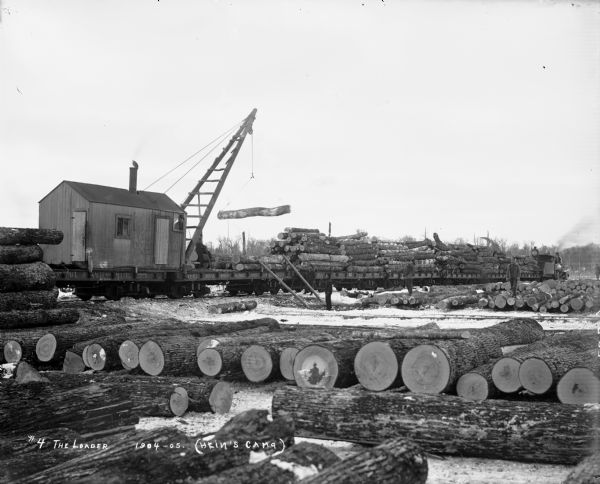 At Hein's logging camp, a crane mounted on a railroad train lifts logs onto a flatbed railway car. Other logs are stacked on the snowy ground. A number of men are working in the background.