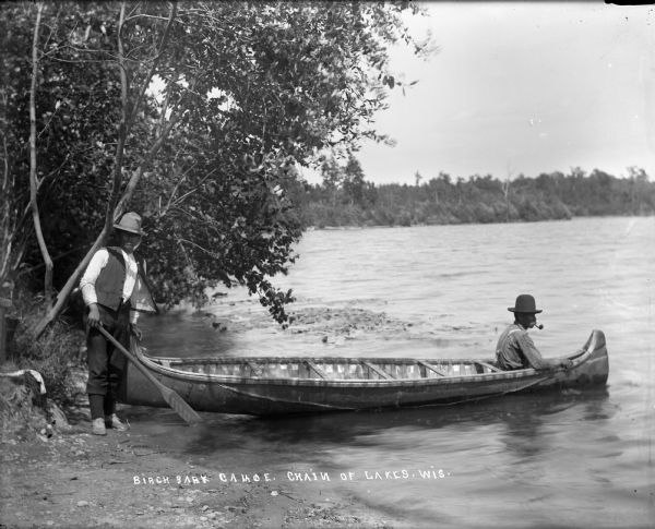 One man sits in the bow and one man stands on shore holding the stern of a birch bark canoe at the edge of a lake. They may be Indians. The man in the canoe is smoking a pipe.