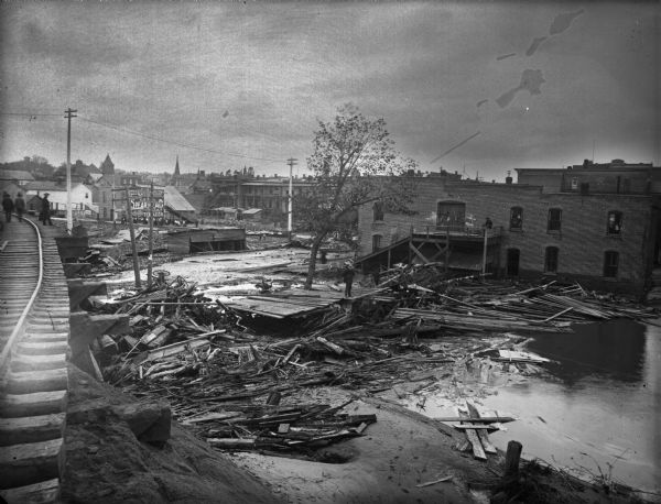 View from railroads tracks of the wreckage of wooden buildings left behind after a flood. Intact buildings are visible in a skyline in the distance, and pedestrians are out looking at the debris.