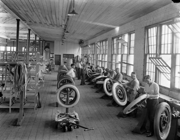Elevated interior view of the Boone Tire Factory. Men are lined up along a wall with open windows. They are each using a stand to hold a wheel while wrapping a layer of canvas or rubber around the outside edge of the tire. Shelves on the right hold canvas and strips of rubber. Several other men stand between racks of tires, observing them. The room also contains machinery and shelving.