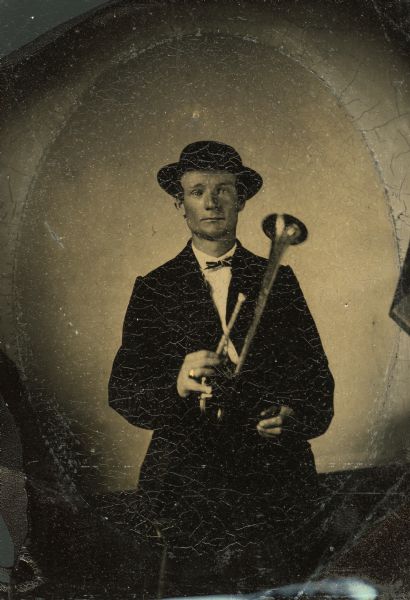 Tintype portrait of John Pickering of Evansville, Wisconsin, member of the 3rd Wisconsin Infantry band. He is standing in a suit and holds a B-flat horn. His wears a ring that is hand-colored gold.