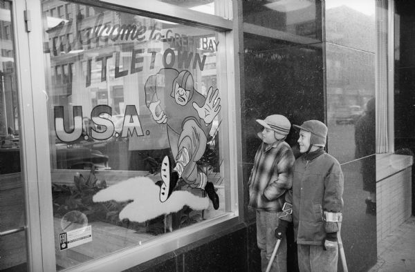 Two young boys, one with Loftstrand-style crutches most likely due to polio, are looking at a window display. The Packers were about to play for, and would go on to win, their seventh title as World Champions of Pro Football. Text written on the window reads "Welcome to Green Bay, Titletown U.S.A."
