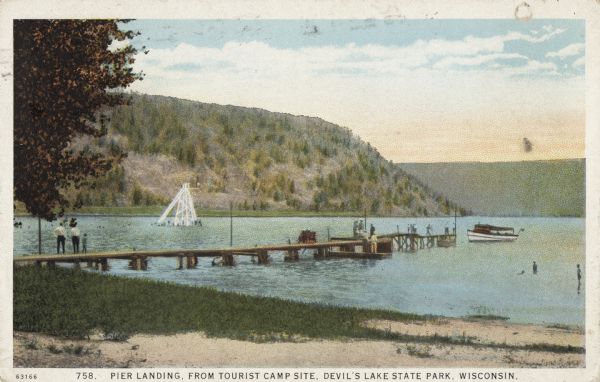 Colorized postcard of the pier landing as seen from the tourist camp site in Devil's Lake State Park. People are strolling on the dock, a boat is docked, with another boat just off the end. Beach and grass are in the foreground. Beyond the dock is the diving tower near the bathing beach. Bluffs, trees and sky are in the background. The text above reads: "Pier Landing, From Tourist Campsite, Devil's Lake State Park, Wisconsin."


