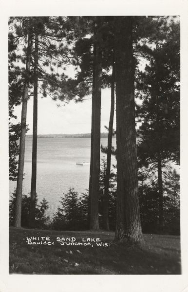 Photographic postcard from hill of White Sand Lake seen through trees. A raft with a diving platform is near the shoreline. Caption reads: "White Sand Lake, Boulder Junction, Wis."