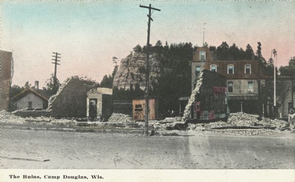 Colorized postcard of "The Ruins." A brick hotel (possibly the MLAKAR HOTEL) is behind and to the right of the old foundation. Various signs are pasted on one of the walls that is still standing. A bluff is in the background. Caption reads: "The Ruins, Camp Douglas, Wis."