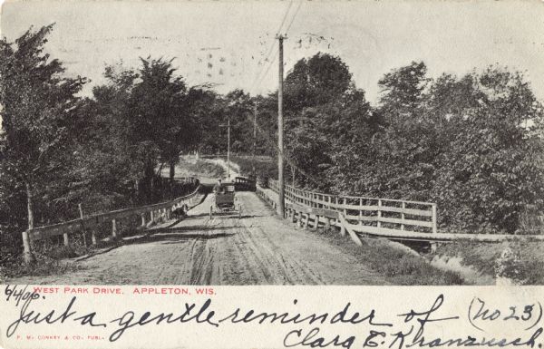 View of West Park Drive. Wooden railings line the road and a wooden walkway is on the right. A man wearing a cap is driving an automobile across the drive. Caption reads: "West Park Drive, Appleton, Wis."