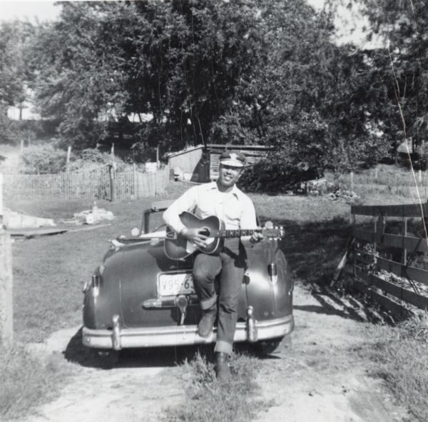 Lewis Arms sits on the trunk of a 1949 convertible (DeSoto?) automobile, strumming a guitar.