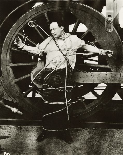 Harry Houdini in chains and ropes. He is tied to a wheel on a locomotive.