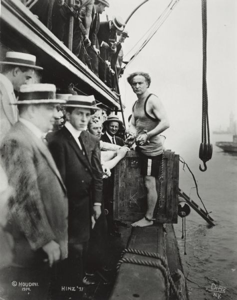 Harry Houdini, born Erik Weisz in Hungary and raised as Erich Weiss in Appleton, Wisconsin, preparing to do one of his famous escapes, this time from a submerged underwater crate. He is standing on the rail of a boat with a crowd surrounding him.