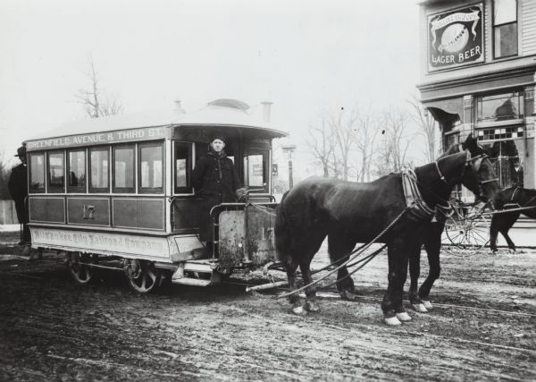 Man driving a horse-drawn trolley of the Milwaukee City Railroad Company on the Greenfield Avenue & Third Street line. The corner of a building advertising Schlitz beer is in the background. Another man stands at the back of the trolley.