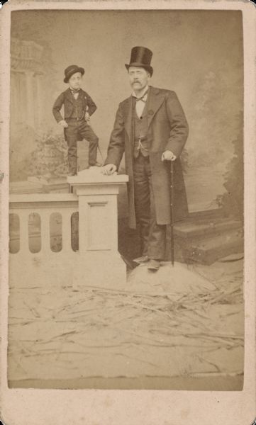 Carte-de-visite of Major Tot standing on a prop column and wall next to a man of average height in front of a painted backdrop. Both men are holding canes. Major Tot's actual name was Pierre Albert Poitras, born in Fitchburg, Massachusetts in 1869. He was hired by P.T. Barnum and billed as "The world's smallest perfectly developed man." He eventually grew to a height of 3.5 feet, weighed ninety pounds and wore a child's size 5 shoe. His father is said to have transported him to engagements in a 14" satchel. Text on the back reads, "Big Success Everywhere! Of Major Tot, The Wonderful Human Midget! Age 16 Yrs, Weight Only 10.5 Lbs."