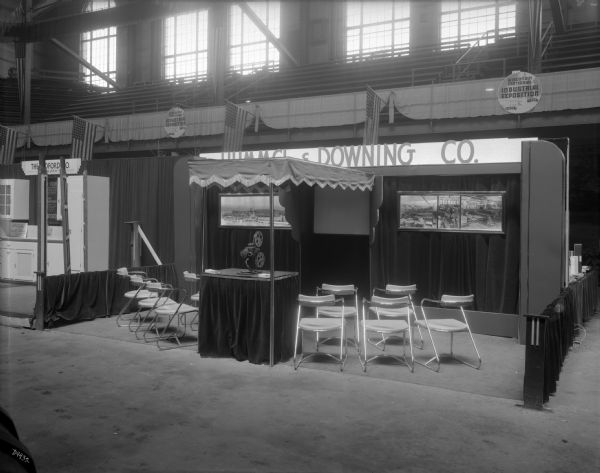The Hummel & Downing booth at the Wisconsin Centennial Industrial Exposition held in the University of Wisconsin-Madison Field House. Hummel & Downing were manufacturers of fibre and corrugated paper and paper boxes. A small projection screen and film projector are set up under an awning flanked by two panoramic images of exterior and interior images of the factory.