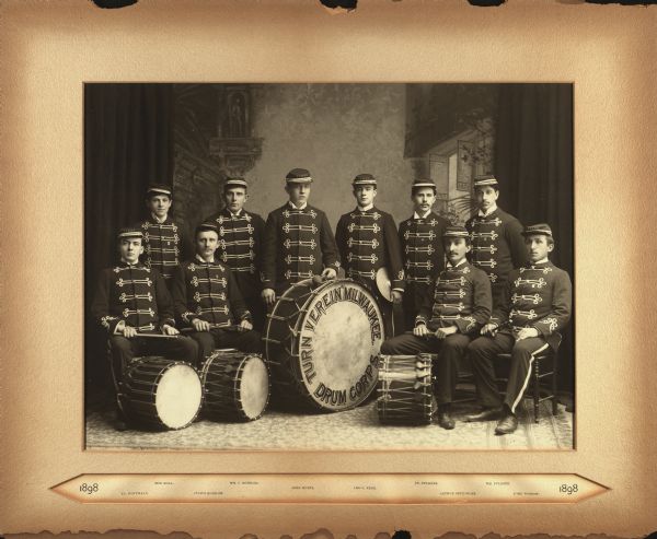 The ten members of the Drum Corps of the Milwaukee Turners posed in front of a painted backdrop. They are dressed in band uniforms with hats and are holding drumsticks. One bass drum and three smaller drums sit on the floor at their feet. On the bass drum is the text, "Turn Verein Milwaukee Drum Corps."