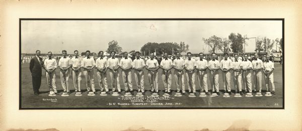 Men from the Milwaukee Turners, lined up outside in a field at the 31st National Gymnastics Festival in Denver. They are in uniform, holding canes(?) with their hats at their feet. A man in a suit stands on the left.