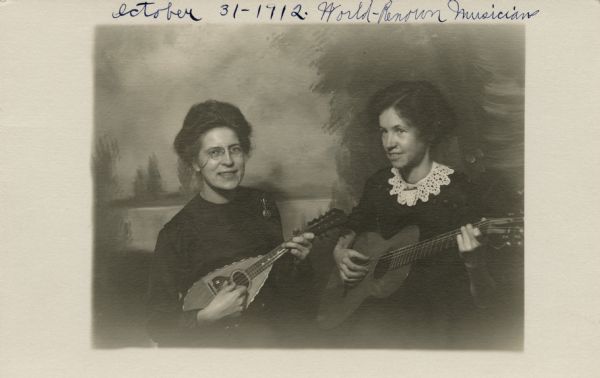 Photographic postcard of two sisters posed with a guitar and a mandolin in front of a painted backdrop. They are wearing dresses, and one sister has a lace collar. At the top of the postcard is written, "October 31-1912. World-Renown [sic] Musicians." On the back is the note, "Dionne sisters, Milwaukee (?), ca. 1912."