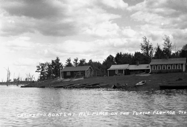 View from water of vacation cabins and boats along lake shoreline of Turtle Flowage. The roof of the building on the right says: "Al's Place / Lunches — Cabins — Beer."