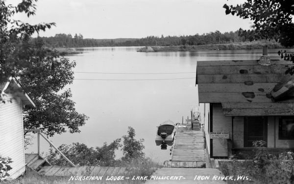 View looking out over Lake Millicent from steep hill at Norseman Lodge. In the foreground is a chair and diving board on the dock. One boat is tied to the dock and has a Caille boat motor. There are two buildings visible, the one on the right has a sign that says “Motor Boat Service, Lake Trips – 50c per person, Towing - $1.00.”
