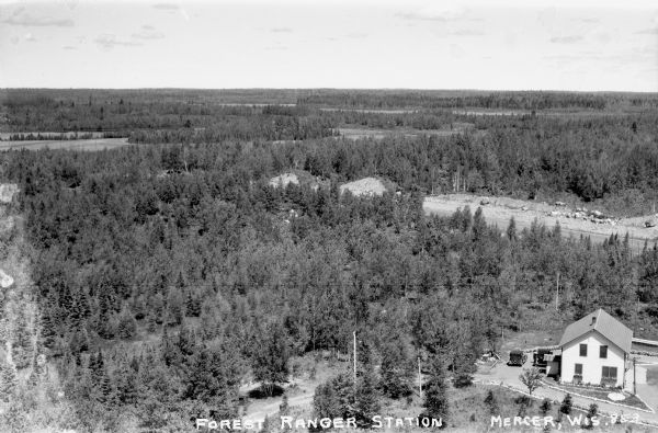 Elevated view of the forest and the two-story Forest Ranger Station with two automobiles parked out front.