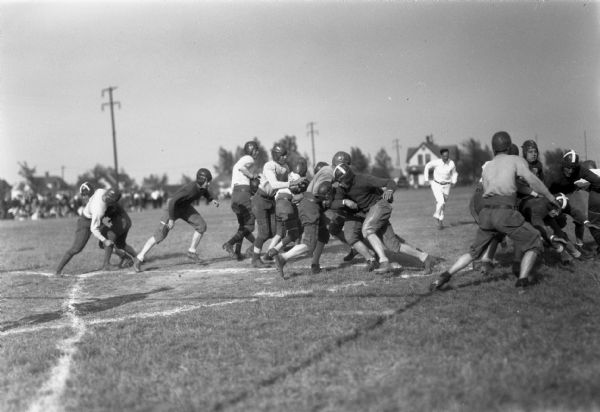 Men wearing helmets tackling each other on a field during a football game. A man in white (referee) is running toward the camera. In the distance is a crowd of people, utility poles, and houses.