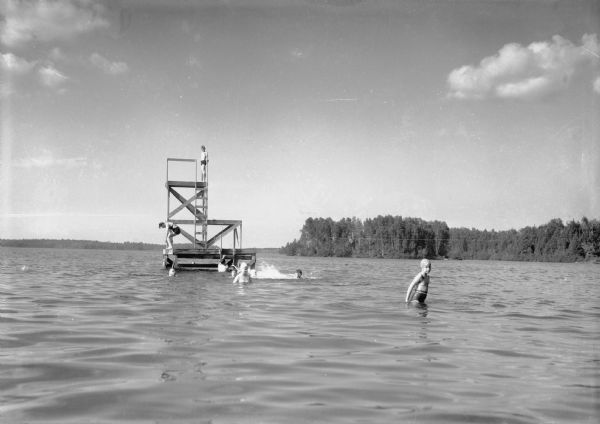 View of men, women, and children wearing swimsuits playing on and swimming near a wooden swimming and diving platform in a lake. In the distance is the far shoreline.