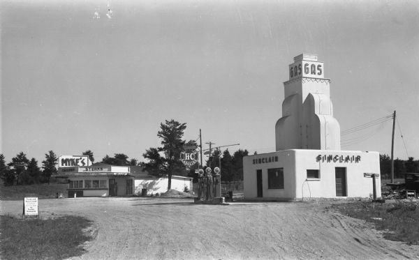 Myke's and Sinclair Gas Station View of Myke's Drive-In and a Sinclair Gas Station with gasoline pumps. Signs say “Trucks and buses travel 2 billion miles a year on Sinclair lubricants," “Myke’s Chicken, Steaks,” and “HC Sinclair Gasoline.”