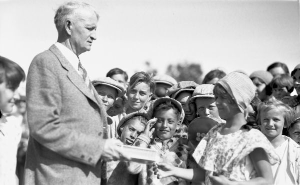 Girl wearing a hat and dress receiving an award from a man wearing a suit and tie while other children in a crowd around them watch.