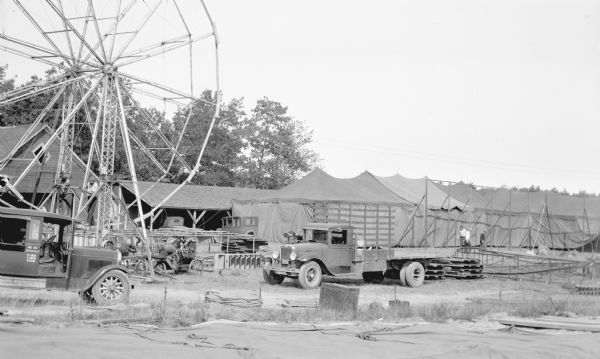 The set up or take down of a Northern Wisconsin County Fair, showing men working on a Ferris wheel and large tents. There is a truck in the foreground and other automobiles visible in the photograph. The automobile on the far left has some numbers by the door that read “No. 22. W.4235, C.2500, T.6735.”