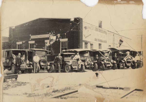 Six trucks and a car are parked in front of the Central Baking Company building. Most of the vehicles are decorated with flags and streamers for a parade. They also have painted sides which advertise Bamby Bread. Men are posed standing next to the vehicles.