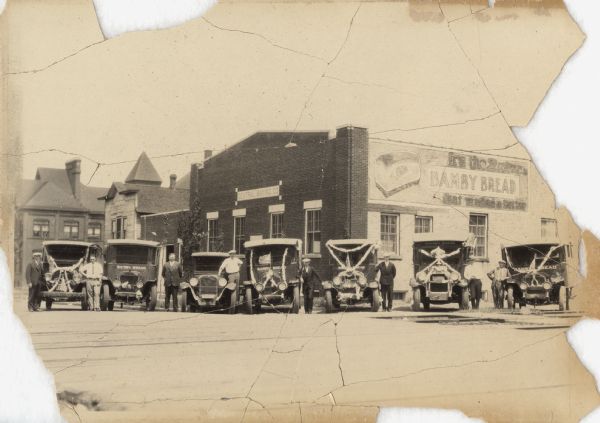 Six trucks and a car are parked in front of the Central Baking Company. Most of the vehicles are decorated with flags and streamers for a parade. The trucks have the words: Bamby Bread" painted on the front; men are posed standing next to the vehicles.