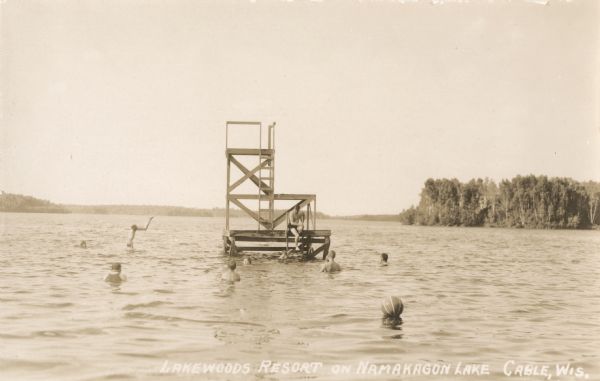 View across water of men and children swimming around and diving off of a diving platform at Lakewoods Resort on Namakagon Lake. In the distance is the far shoreline lined with trees.