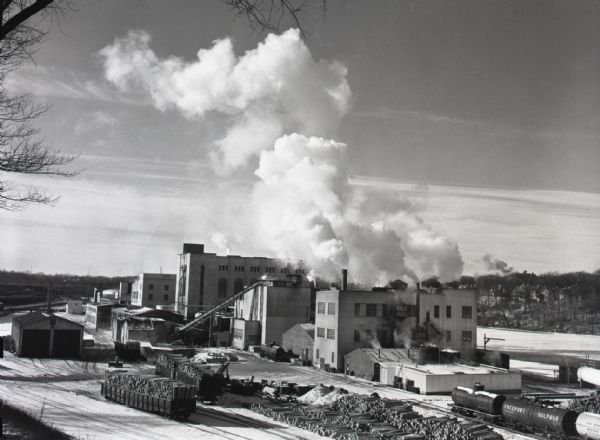 Smoke billowing from smokestacks at the Consolidated Paper plant. The river appears to be partially frozen, and snow is on the ground. Piles of logs, on the ground and in open railroad cars, are in the foreground.