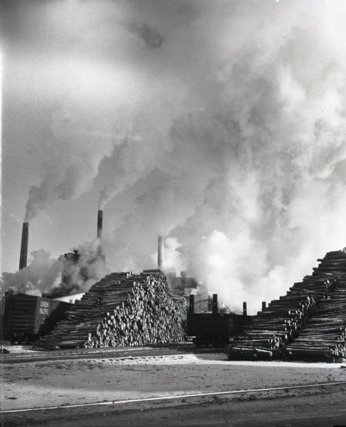 View across railroad tracks of billowing smokestacks at the Thilmany Paper Company plant. Stacks of logs are piled next to railroad tracks and cars.