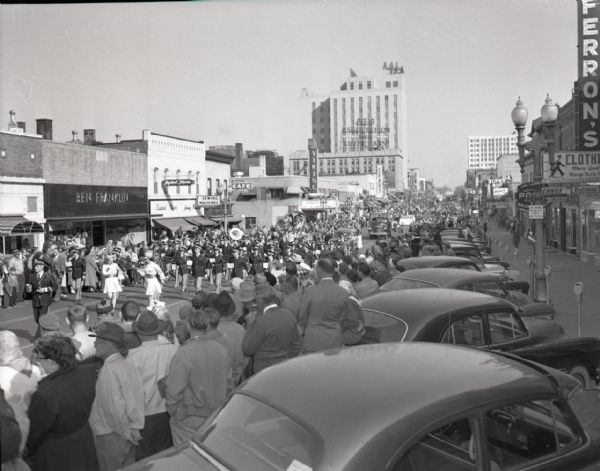 Street view of a parade on College Avenue at Walnut Street. Crowds and cars line both sides of the street, looking on as bands, cars, and floats pass by. Local businesses are also visible, including a Ben Franklin store.