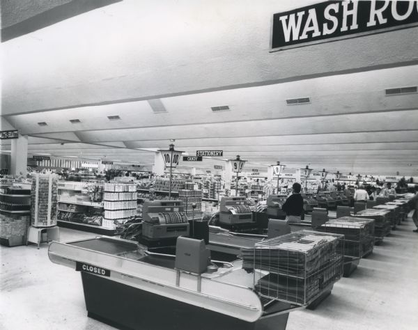 Interior of the Treasure Island Store, located on West College Avenue. Store aisles and end-caps full of goods are visible. A single cashier waits in aisle 8 while other cashiers assist customers in the background. Each check-out counter has a stack of newspapers at the end. The Treasure Island was the first discount box store in the Fox River Valley.