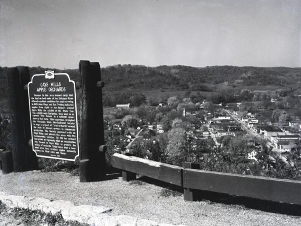 Image of the historical marker for Gays Mills Apple Orchards overlooking the town of Gays Mills, Wisconsin. The marker stands to the left, leaving an elevated view of the town's business and residential areas. Cars are lining the street along one of the main roads.