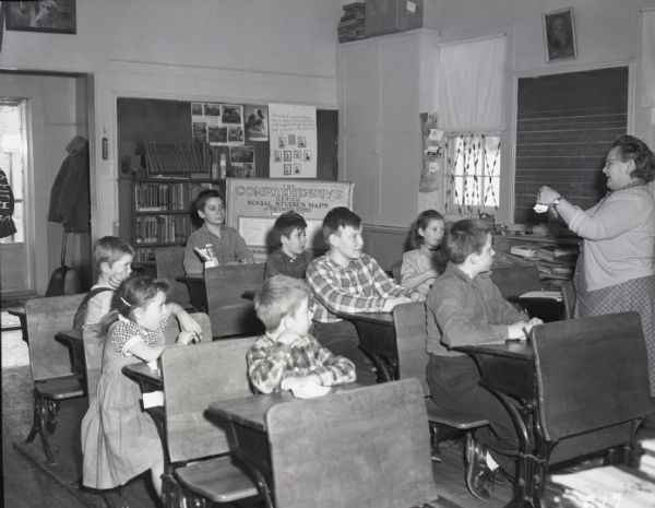 Class of young boys and girls in a one-room school house. The children sit in three rows of school desks, watching their teacher as she instructs. Books and educational literature are visible in the background.