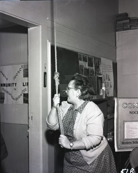 Teacher pulling on a rope to ring the school bell in a one-room school house. School literature and a chalkboard are visible in the background.