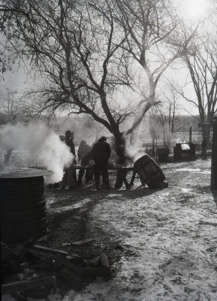 Far view of farmers preparing a hog for slaughter. A group of farmers stand around a hog on a long table outside. Smoke and steam billow from a stove in the foreground and wooden barrel leaning against the table.