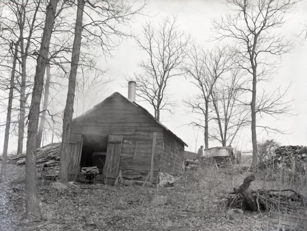View of a maple syrup shack on the side of a hill. Smoke is billowing out of the doorway, which is blocked by a wheel barrel full of wood. Two men are in the background next to a trailer and a tractor, and large metal trough.