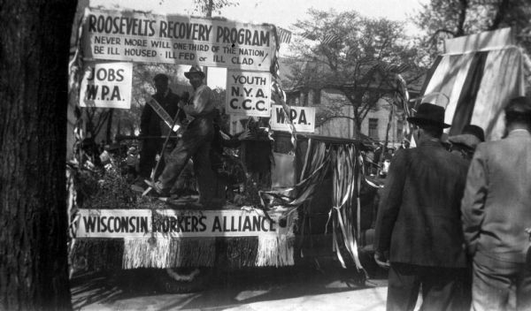 The Wisconsin Worker's Alliance May Day parade float. The float has three workers on it with tools and signs read "Roosevelt's Recovery Program: Never More Will One Third of the Nation Be Ill Housed, Ill Fed or Ill Clad!" "Jobs W.P.A." "Youth N.Y.A. C.C.C."