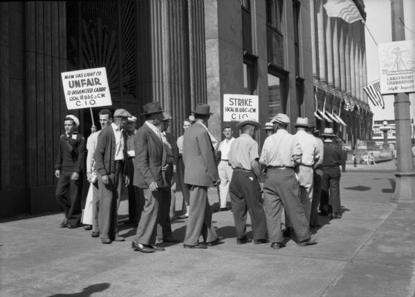 Striking workers from the local 18 of United Gas Coke and Chemical Workers, C.I.O. picketing on a wide downtown sidewalk. The men are walking in a circle holding signs. One reads "Milw. Gas Light Co. UNFAIR to organized labor." A navy seaman passes the picketers on one side. Large buildings with decorative metal work and columns rise in the background.