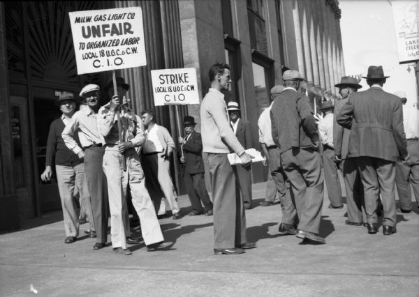 Members of local 18 United Gas, Coke and Chemical of the C.I.O. picketing in a small circle on a wide downtown sidewalk. Two men hold signs and one reads, "Milw. Gas Light Co. UNFAIR to organized labor."  Another sign declares, "STRIKE." A large stone building with decorative metal-work and columns stands in the background.