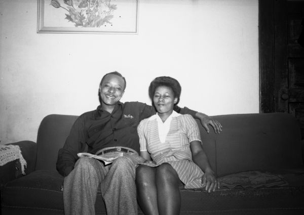 Isaiah Pyant, C.I.O. union member, and his wife sitting on a couch in their home. They are smiling and relaxed and a print of a bouquet hangs on the wall behind them.