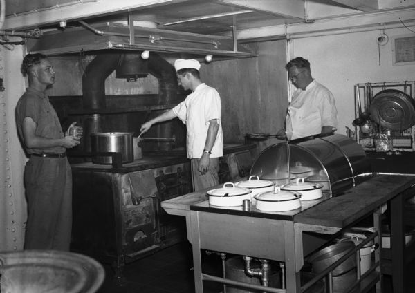 Cooks prepare food on a large iron stove while a worker stands holding coffee. Part of a series on working conditions aboard Lake Michigan railroad car ferries. Ferry workers belonged to the National Maritime Union (NMU).