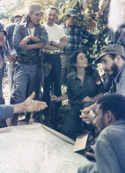 Meeting of the 26th of July Movement in the woods of Oriente Province during the Cuban Revolution. Fidel Castro, Juan Almeida Bosque and Vilma Espin crouching around an unrolled map in the country. A number of men are standing behind them listening.