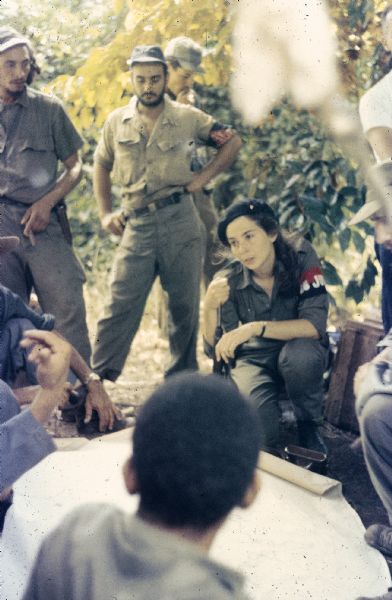 Meeting of the 26th of July Movement in Oriente Province during the Cuban Revolution. A number of participants, including Vilma Espin, Raúl Castro and Juan Almeida Bosque, are sitting, standing and crouching around a map laid on the ground in the countryside.